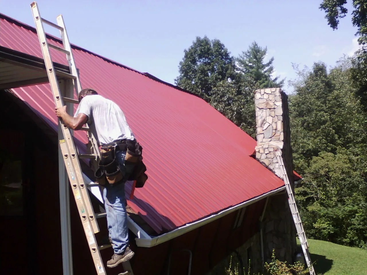 Roofer on a ladder beside a red metal roof and stone chimney — Soffit & Fascia Repair by Roofing Brothers in Mc Queeney